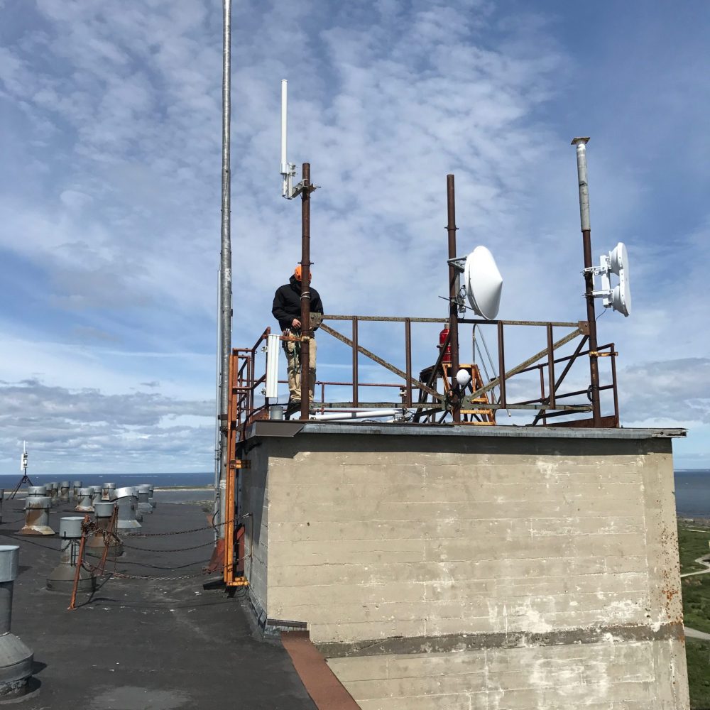 Technicians installing network antennas and point-to-point wireless dishes on a rooftop platform under a partly cloudy sky