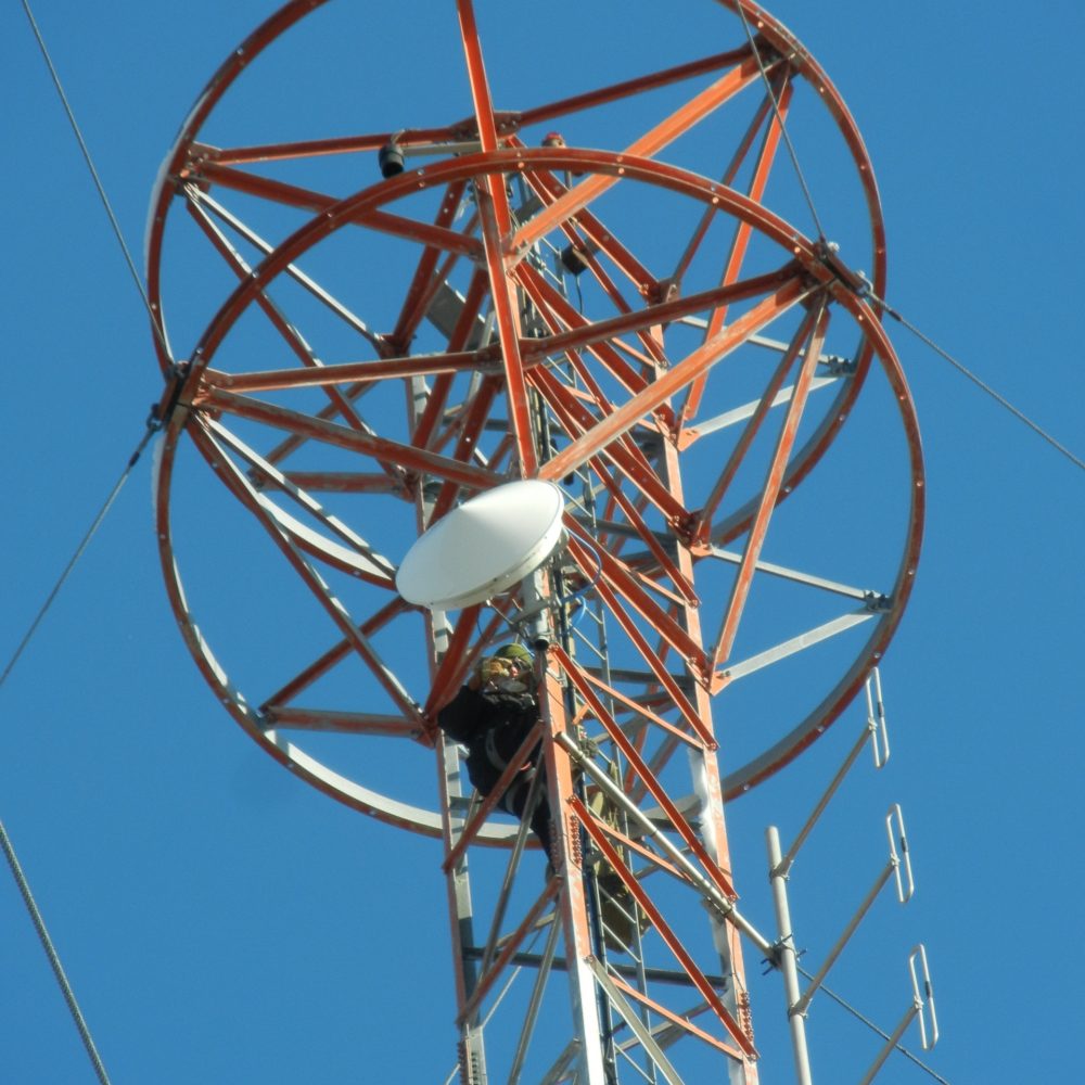 Technician climbing a red and white broadcast tower to install or maintain communication equipment under a clear blue sky