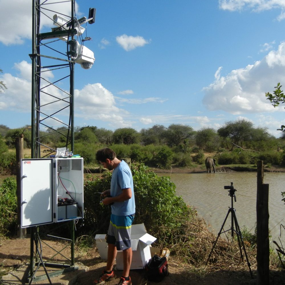 Technician installs a wildlife camera system at a watering hole on Mpala Ranch in Kenya, with an elephant in the background.