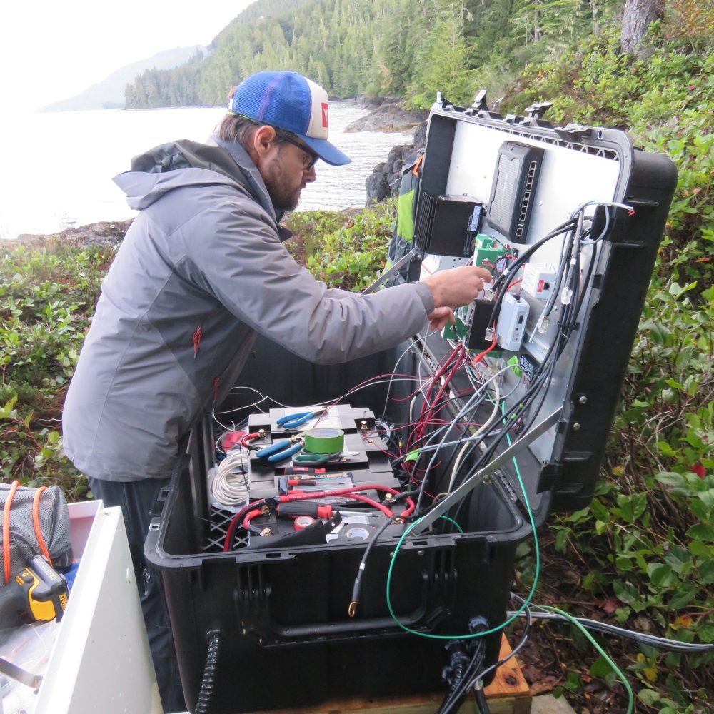 Technician working on off-grid power system with batteries and wiring in a remote coastal forest