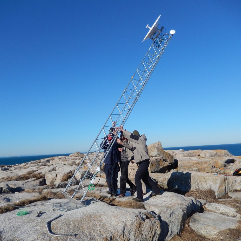 Crew raises a communications tower on a rocky coastal outcrop under a clear blue sky.