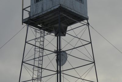 A weathered wooden cabin on top of a tall metal tower with a ladder, satellite dish, and security camera against a cloudy sky.